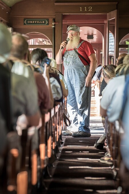 1880 Train narrator in overalls speaking to passengers inside a vintage wooden passenger car labeled Oreville
