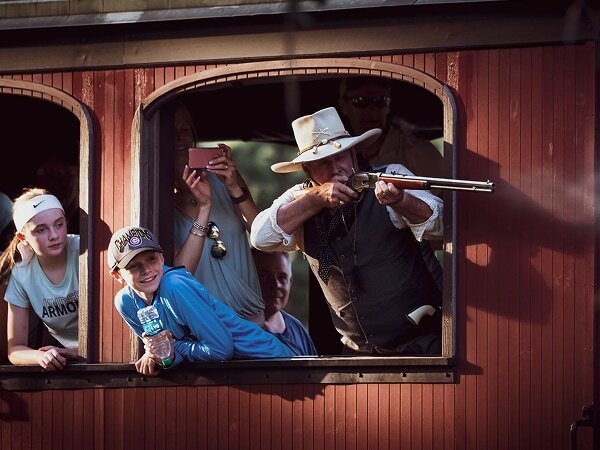 Old West gunfighter performer leaning out of a 1880 Train passenger car window during an Old West Shootout event, with excited kids and passengers watching